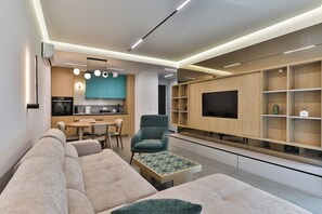 Ein modernes Wohnzimmer mit einer Couch, Sesseln und einem Couchtisch, eine Küchenzeile mit Esstisch und Stühlen sowie ein Fernseher an einer Holzwand.