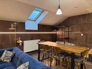 A modern interior with a sloped ceiling, a skylight, a dining area with a wooden table and patterned chairs, and a blue sofa with decorative pillows. - Charming 2-bedroom Apartment in Heart of Prague (Prague)