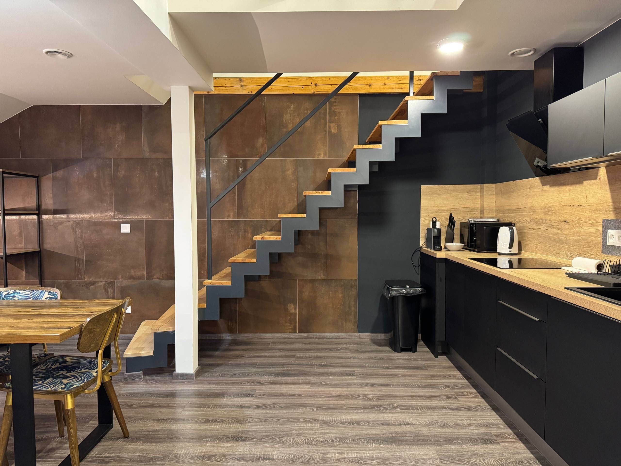 A modern kitchen with a wooden staircase, a dining area with a table and chairs, and a trash bin.