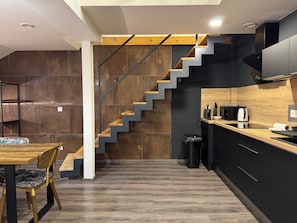 A modern kitchen with a wooden staircase, a dining area with a table and chairs, and a trash bin. - Charming 2-bedroom Apartment in Heart of Prague (Prague)