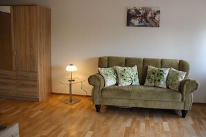 A room with a wooden wardrobe, a green sofa with tree-patterned cushions, a floor lamp, and a painting on the wall. - Holiday apartment with 1 bedroom (Gössweinstein)