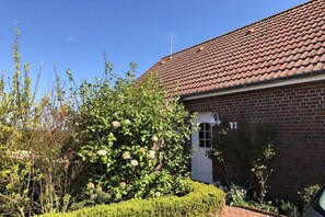A brick house with a red-tiled roof, a white door, and a garden with various plants and a flowering bush. - Friesenhaus RoseCottage in Greetsiel (Krummhörn Greetsiel)