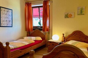A room with two wooden beds, a window with orange curtains, a wall-mounted light, and two small framed pictures. - Apple blossom at the Bayer house (Pottenstein)