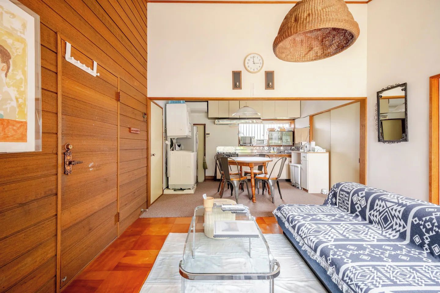A compact living space with a wooden door, a dining area with a table and chairs, a sofa with patterned cushions, and a kitchen area with appliances.