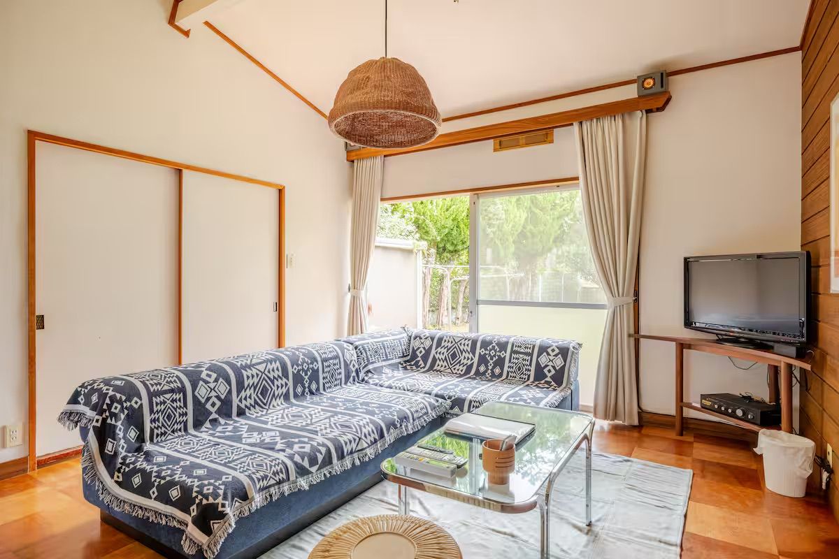 Una sala de estar con un sofá, una mesa de centro de vidrio, un televisor sobre un mueble bajo y un ventanal con cortinas.