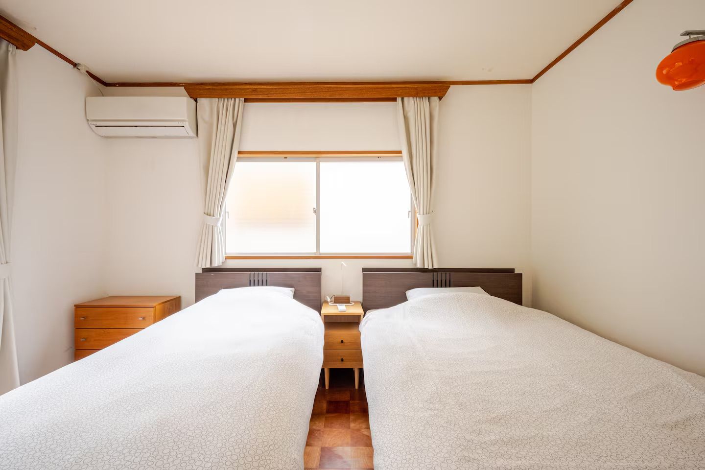 A room with two beds, a window with curtains, a nightstand, and an air conditioning unit.