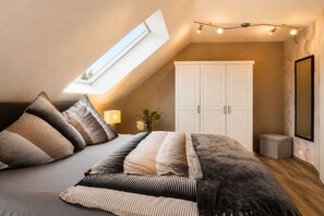 Un dormitorio con techo inclinado, una cama con muchos cojines, un armario, una mesita de noche con una lámpara y un espejo en la pared.