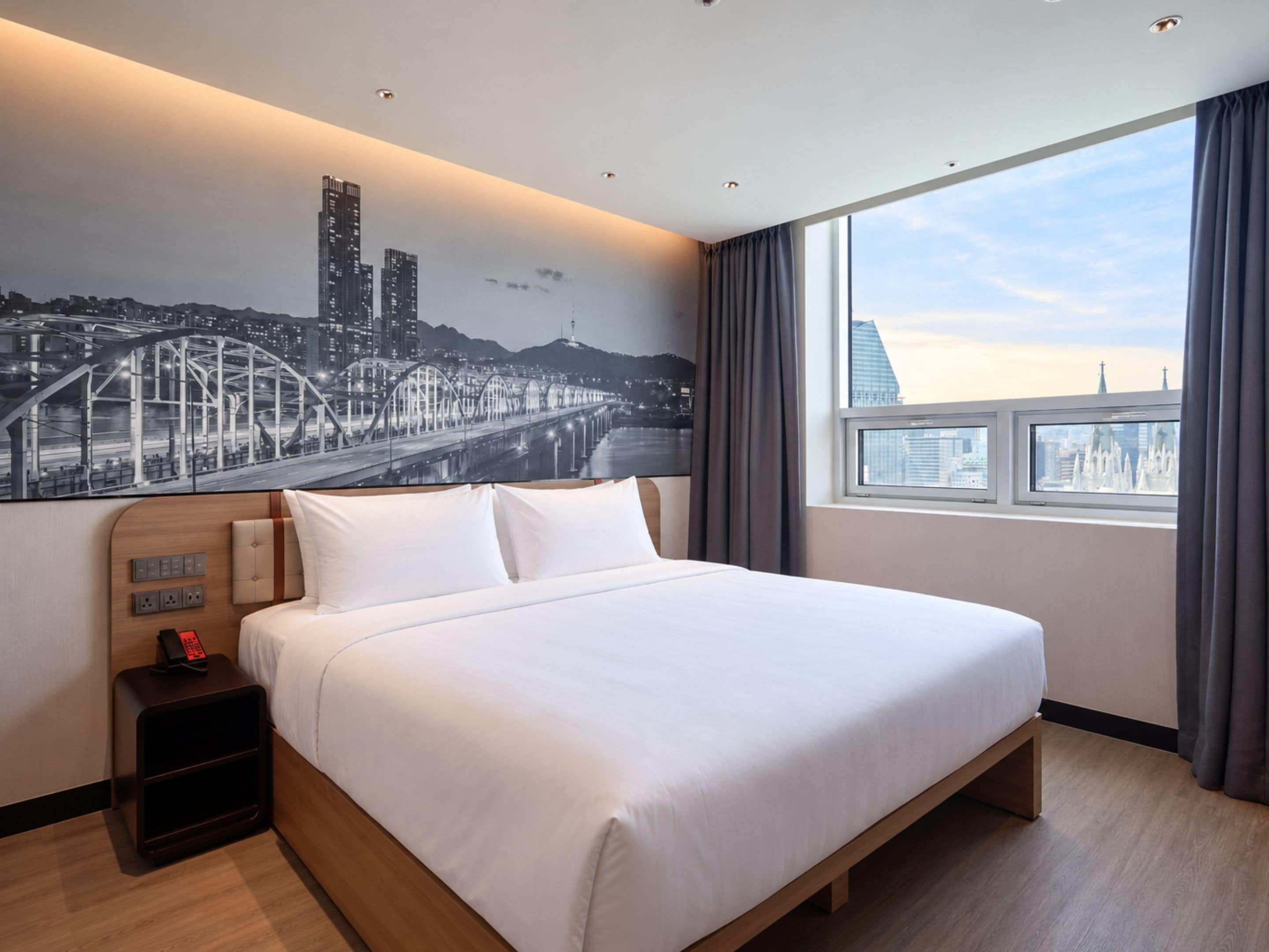 A hotel room with a large bed, a nightstand with a phone, a wall-mounted light switch, and a window with a cityscape view.