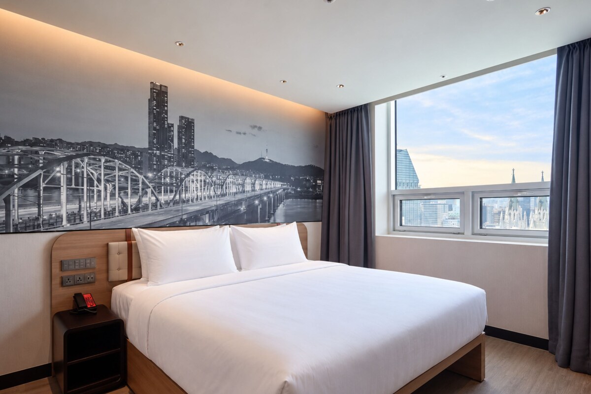 A hotel room with a large bed, a nightstand with a phone, a wall-mounted light switch, and a window with a cityscape view.
