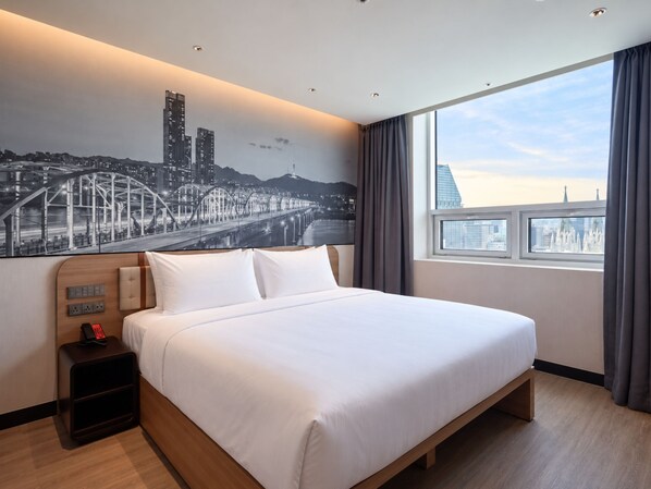 A hotel room with a large bed, a nightstand with a phone, a wall-mounted light switch, and a window with a cityscape view.