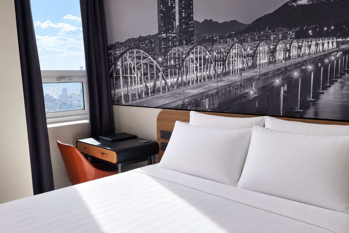 A hotel room with a bed, a desk, a chair, and a large cityscape mural on the wall.