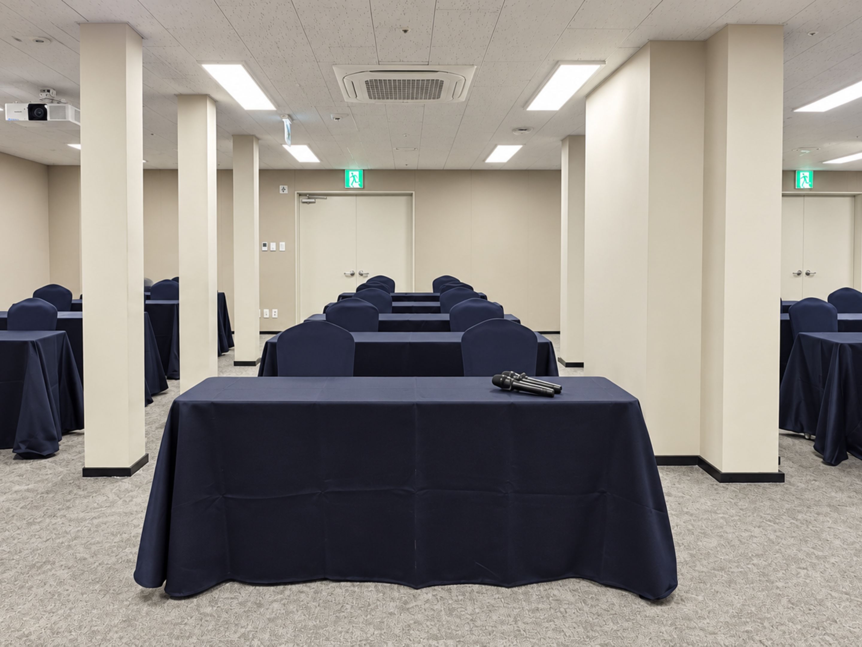 A conference room with rows of tables and chairs, a projector, and an exit sign.