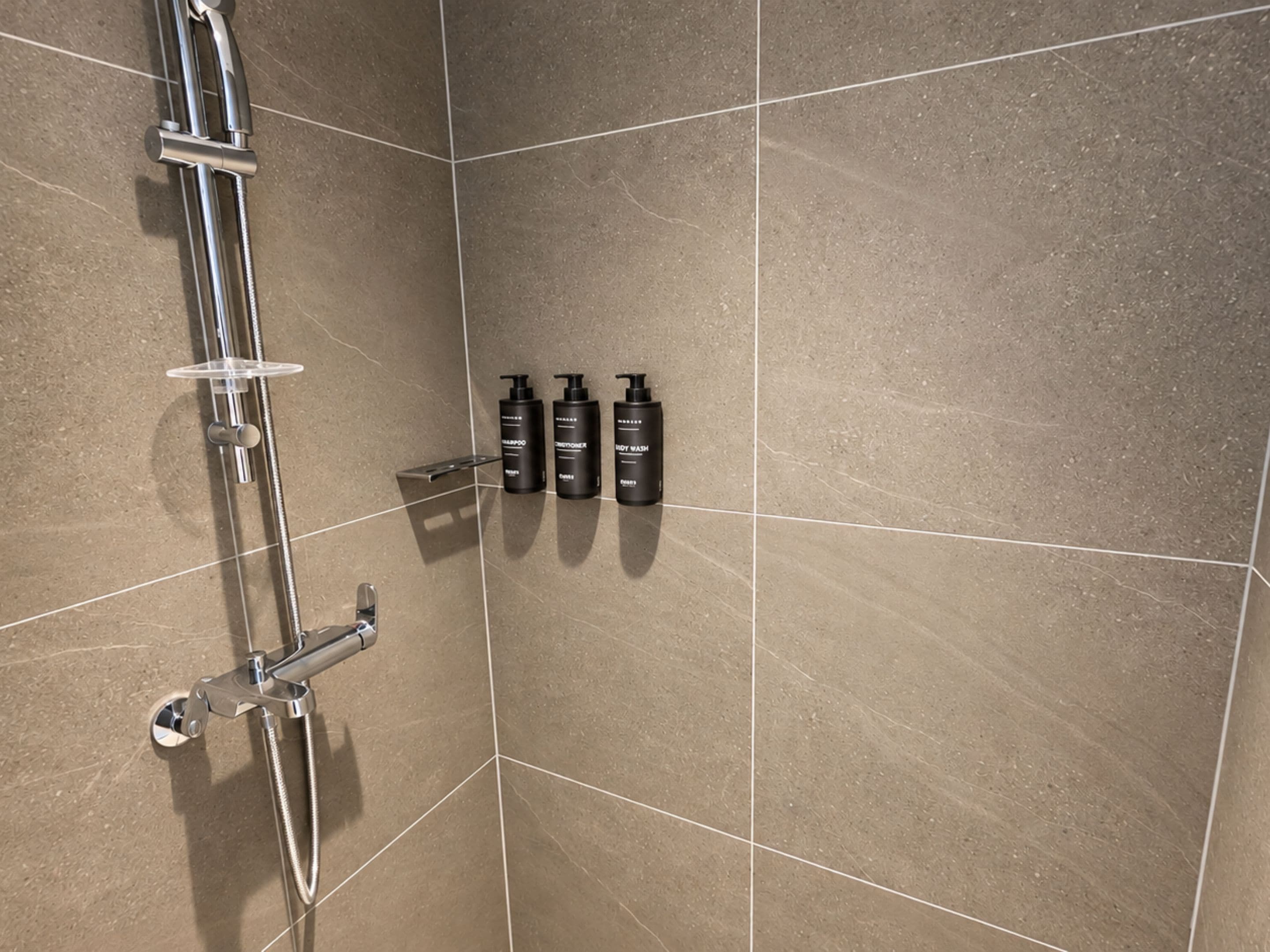 A shower area with a chrome showerhead, a handheld shower attachment, and four bottles of shampoo and conditioner on the wall.