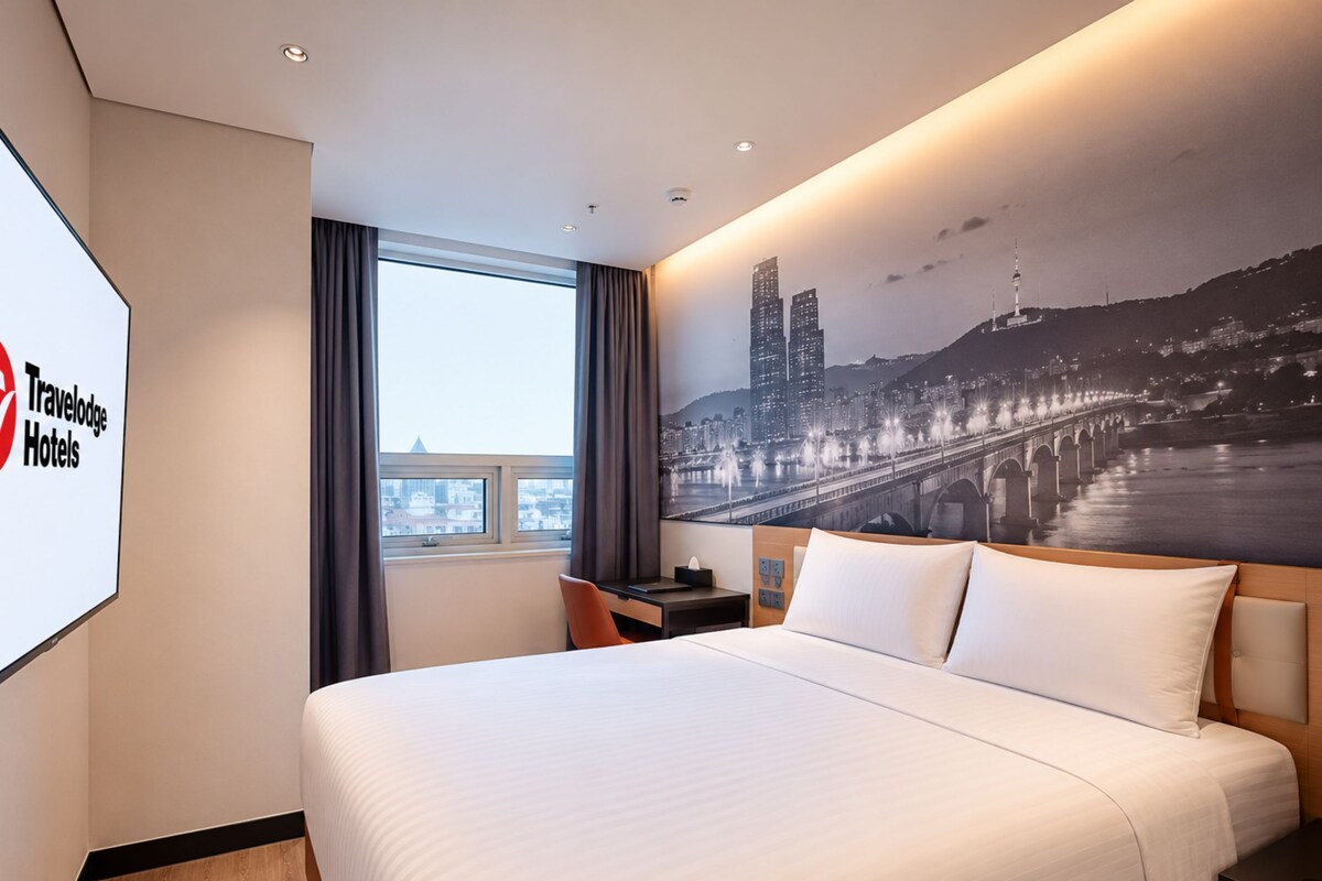 A hotel room with a bed, a desk, a chair, a nightstand, a window, and a cityscape mural on the wall.