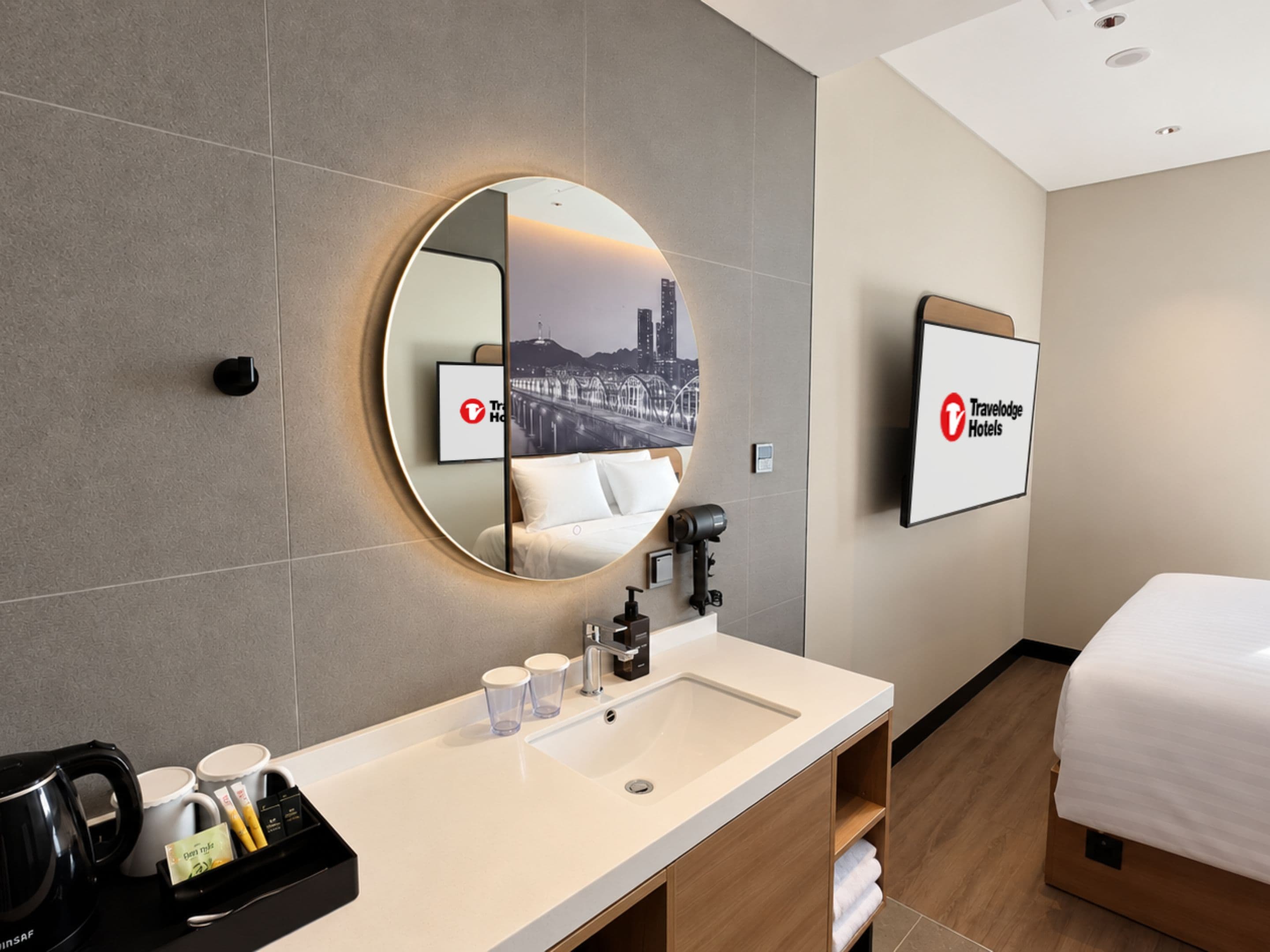 A hotel room with a round mirror, a bed, a sink with toiletries, and a framed picture of a cityscape.