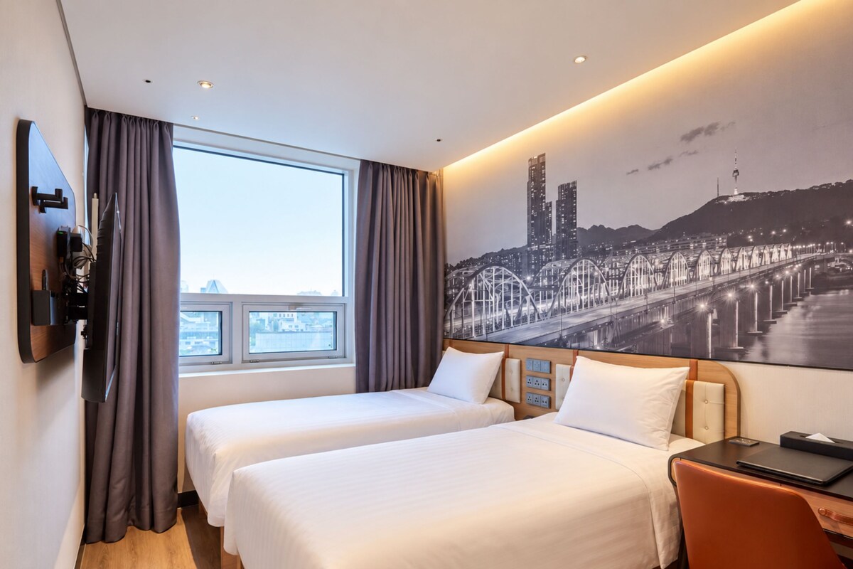A hotel room with two beds, a desk, a TV, and a cityscape mural on the wall.