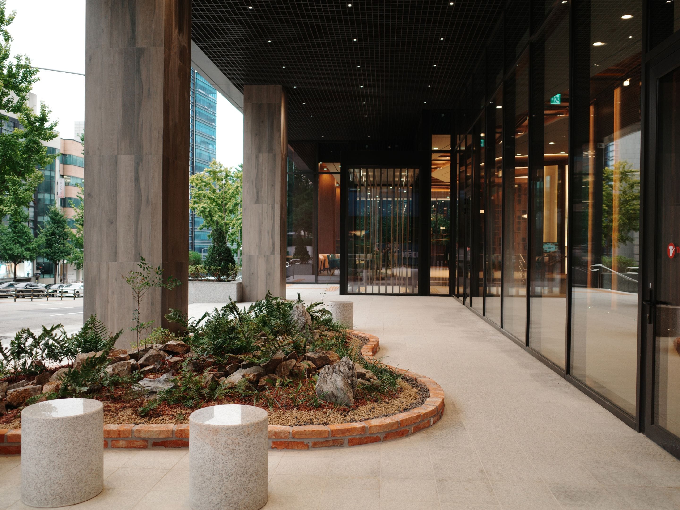 A modern building with a covered walkway, large windows, and a landscaped area with plants and a stone feature.