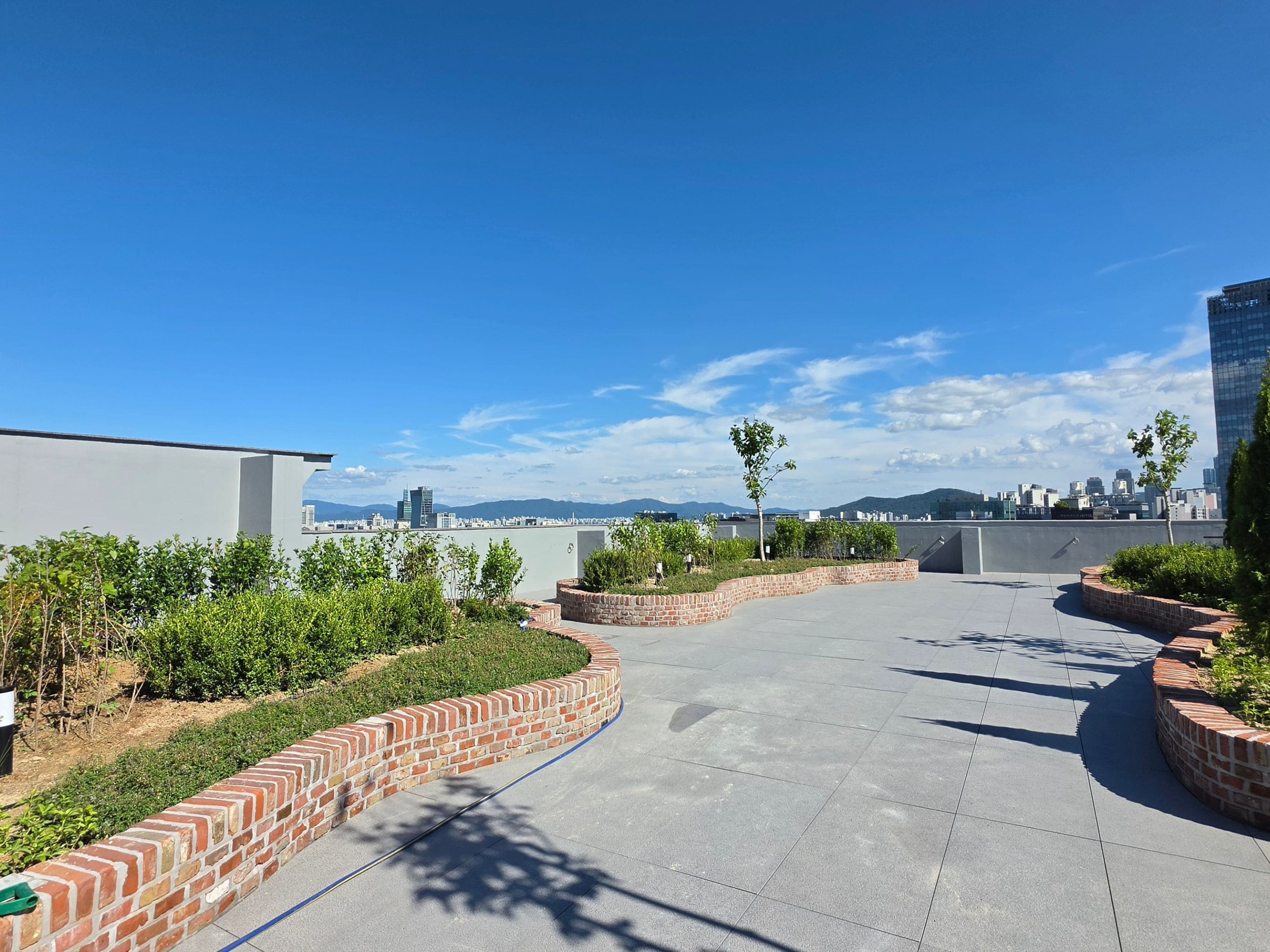 A rooftop terrace with a curved brick pathway, greenery, and a city skyline in the background.