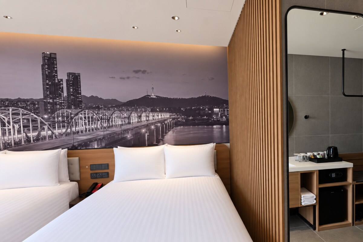 A hotel room with a large bed, a wooden headboard, a mirror, a nightstand, and a cityscape mural on the wall.