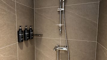 A shower area with a handheld showerhead, a wall-mounted faucet, and a handheld shower attachment. There are three bottles of shampoo and conditioner on a shelf.