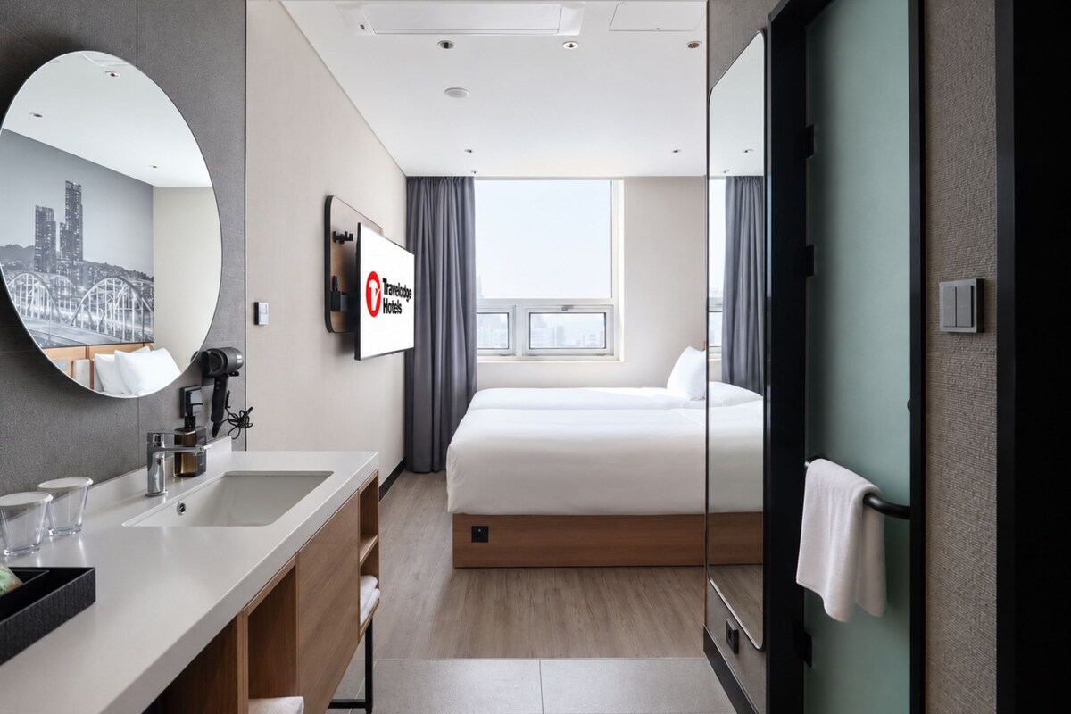 A hotel room with a large bed, a bedside table, a mirror, a TV, and a window with curtains.