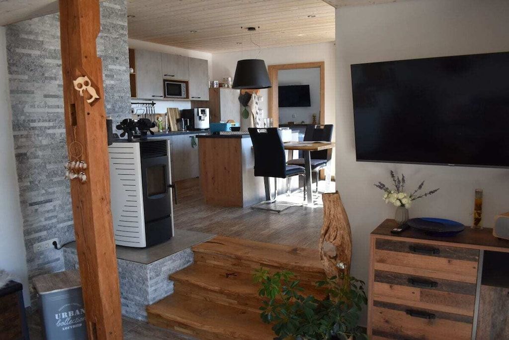 A modern kitchen with a central island, a dining area with a black chair, and a flat-screen TV mounted on the wall.