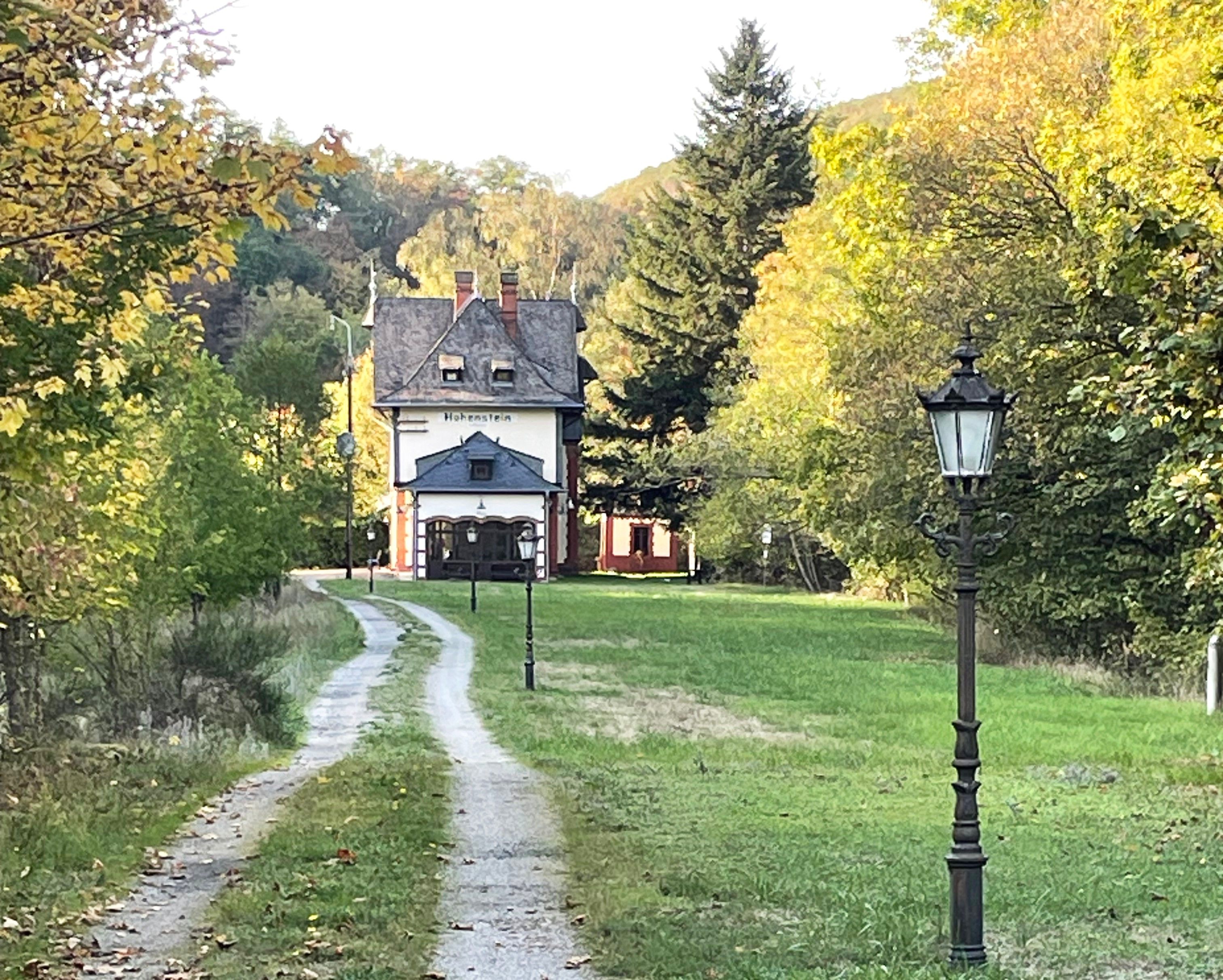 A rural pathway leading to a traditional building with a sloped roof and a sign reading 'Hohenstein'.