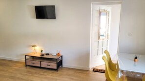 A room with a TV mounted on the wall, a desk with a lamp, a chair, and a small table with a plant. - Homerez - Studio Chauny (Chauny)