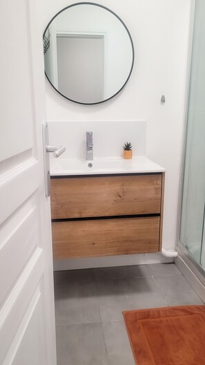 A bathroom with a white vanity, a wooden drawer, a small potted plant, and a glass shower enclosure. - Homerez - Studio Chauny (Chauny)
