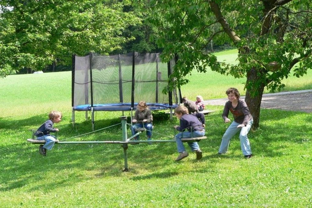 Une aire de jeux avec une balançoire et un trampoline, entourée d’arbres et d’herbe.