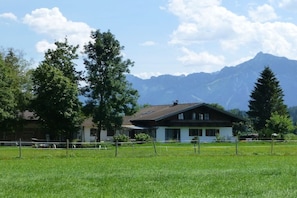 A two-story house with a sloped roof, surrounded by a fenced field and trees, with mountains in the background. - Holiday apartment with 3 bedrooms (Füssen)