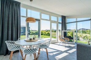 A modern dining area with a round table, white chairs, and a view of greenery through large windows. - Holiday apartment 66 (Ellemeet)