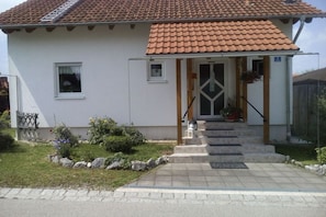 A two-story house with a tiled roof, a front porch with steps, and a small garden with rocks and plants. - Holiday in the Ostallgäu region (Lengenwang)