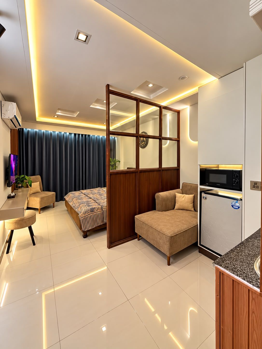 Une chambre d’hôtel moderne dotée d’une kitchenette, d’un lit avec une literie à motifs, d’un coin salon avec un canapé et un fauteuil, et d’une grande fenêtre avec des rideaux.