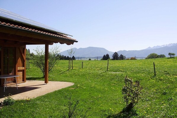 Una cabaña de madera con un patio, rodeada por un campo verde exuberante y montañas al fondo.