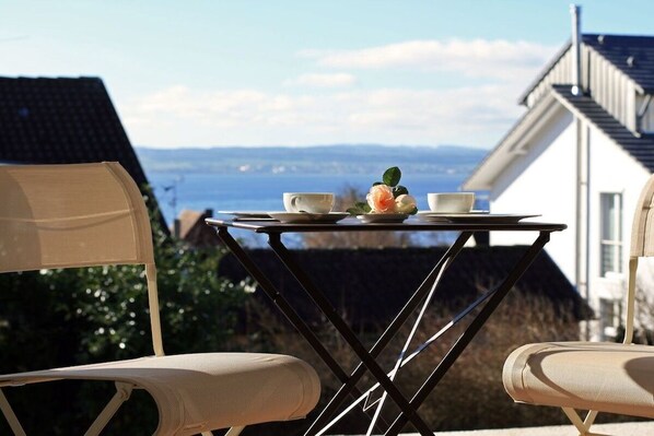 A table with two chairs, a cup, and a plate with a flower, set against a view of a house and landscape. - "Lake in view" in Hagnau on Lake Constance (Hagnau)