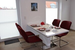 A dining area with a white table, four red chairs, and a window with blinds. - Greetsieler Sonneneck (Greetsiel)