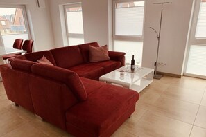 A red sofa, a white coffee table, and a floor lamp in a living room with large windows. - Greetsieler Sonneneck (Greetsiel)