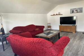 A living room with a red sofa, a coffee table, a TV on a wooden stand, and a small side table with a lamp. - Eifelblick (Apartment Talblick) (Weinsheim)
