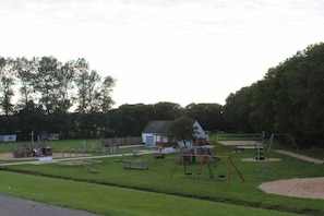 Ein Spielplatz mit Schaukeln und einer Rutsche, umgeben von einer Rasenfläche und Bäumen.