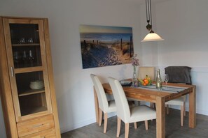 A dining area with a wooden table, white chairs, a cabinet with glass doors, and a painting of a beach scene on the wall. - Holiday apartment with 2 bedrooms (Schönhagen)