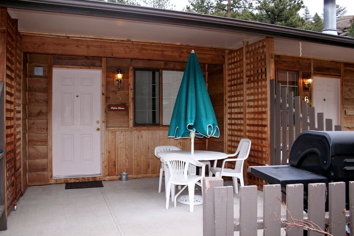 A wooden cabin with a white door, a table with chairs, a barbecue grill, and a green umbrella.