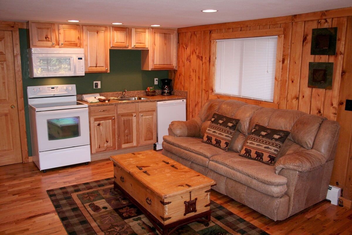 A compact kitchen with wooden cabinets, a microwave, stove, and a refrigerator. There's a sofa with decorative pillows and a wooden chest in the living area.