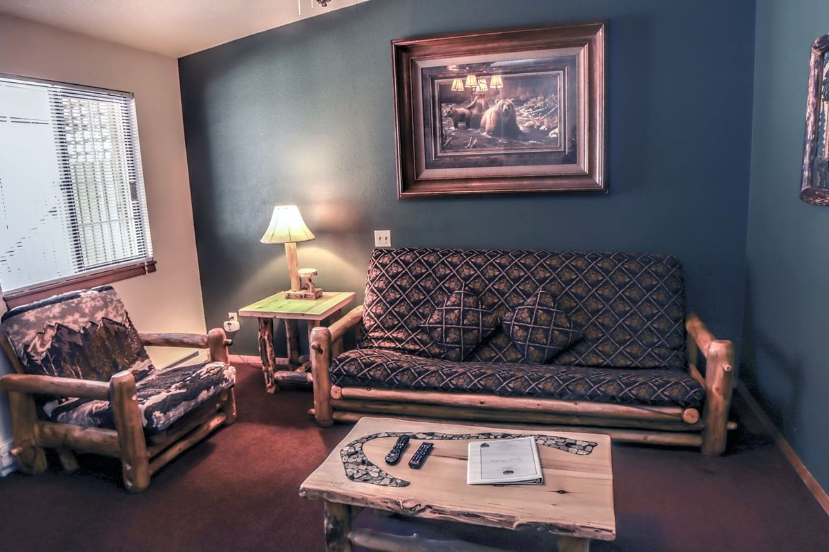 A room with a patterned sofa, a wooden chair, a small table with remote controls and a book, a lamp, and a framed picture on the wall.