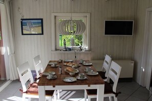 A dining area with a wooden table set for four, surrounded by white chairs. A window with a view of greenery is above the table, and a television is mounted on the wall to the right. - Holiday home House 105 (Damp)
