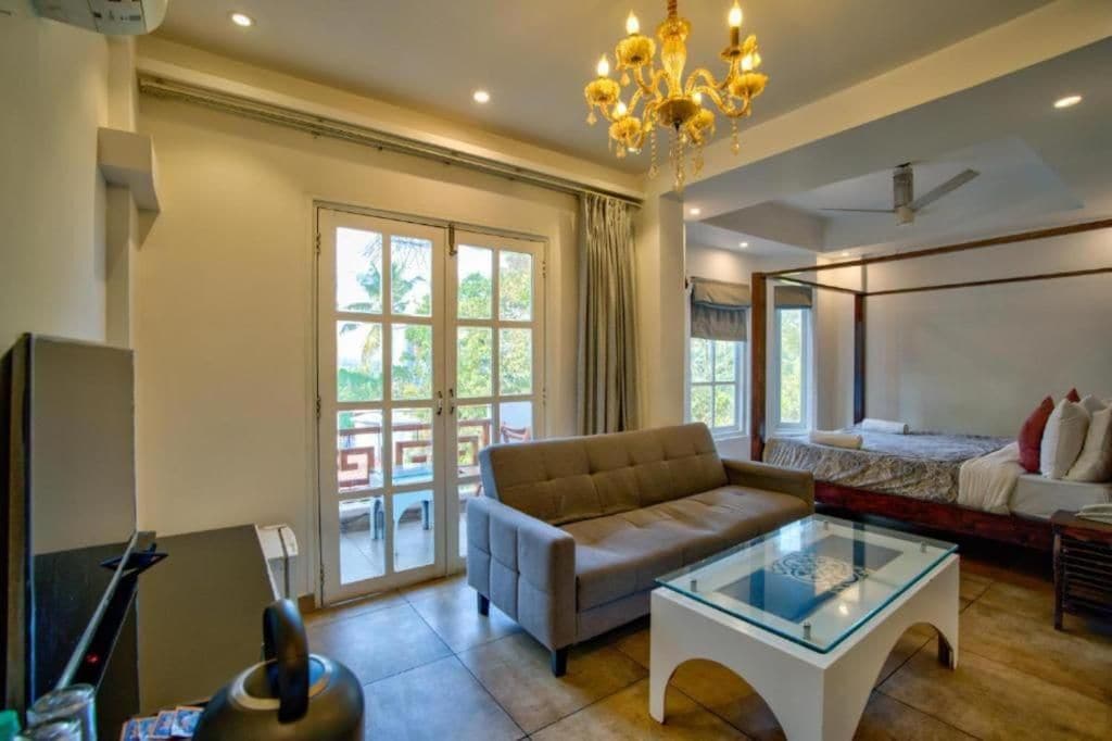 Habitación de hotel con sofá, una mesa de centro de vidrio, cama con almohadas, ventilador de techo y candelabro.