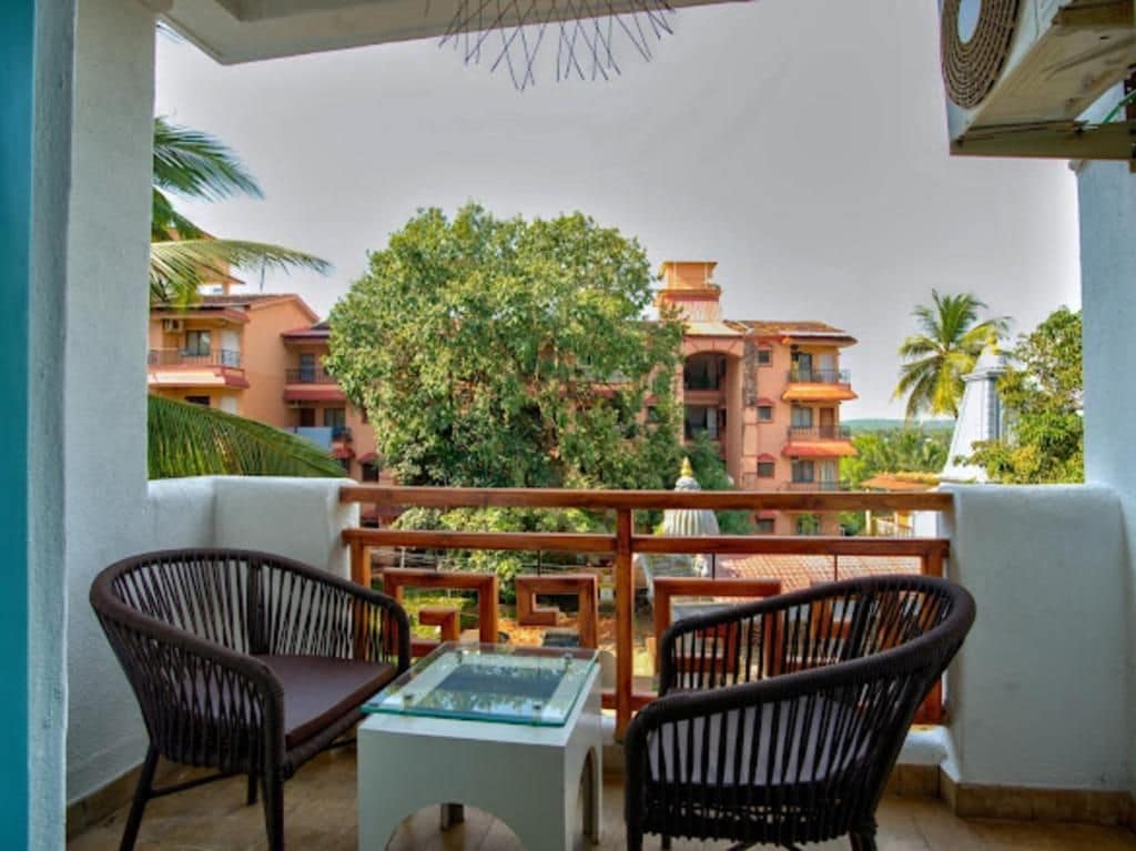 A balcony with wicker chairs and a glass table, overlooking a residential area with buildings and trees.