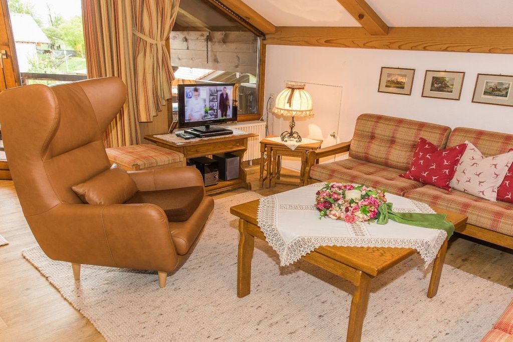 Uma sala de estar acolhedora com poltrona de couro, mesa de centro de madeira com um arranjo floral, sofá xadrez e uma televisão em suporte de madeira.