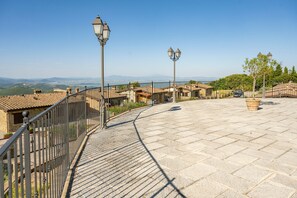 Eine gepflasterte Terrasse mit Metallgeländer, Straßenlaternen und Blick auf entfernte Gebäude und Hügel.