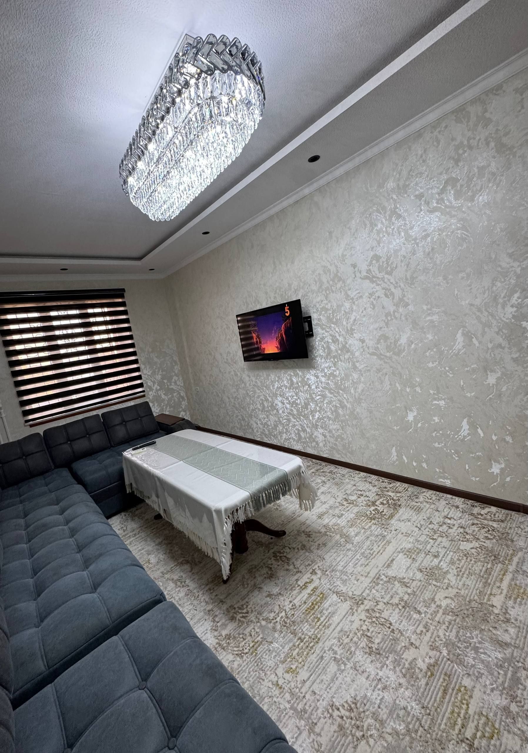 A modern living room with a sectional sofa, a flat-screen TV mounted on the wall, and a decorative chandelier.